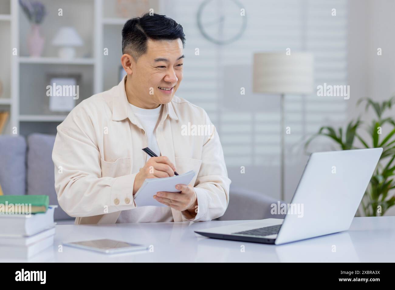 Asiatischer Mann, der sich während eines Online-Webinars zu Hause Notizen macht, mit einem Laptop. Pädagogisches und professionelles Umfeld. Stockfoto