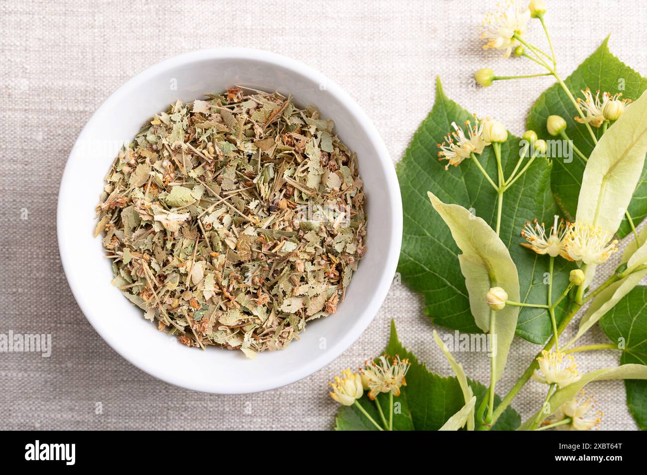 Lindenblumen in weißer Schüssel auf Leinenstoff. Getrocknete und frische Blüten, Früchte und Blätter der großblättrigen Linde oder Linde, Tilia platyphyllos Stockfoto