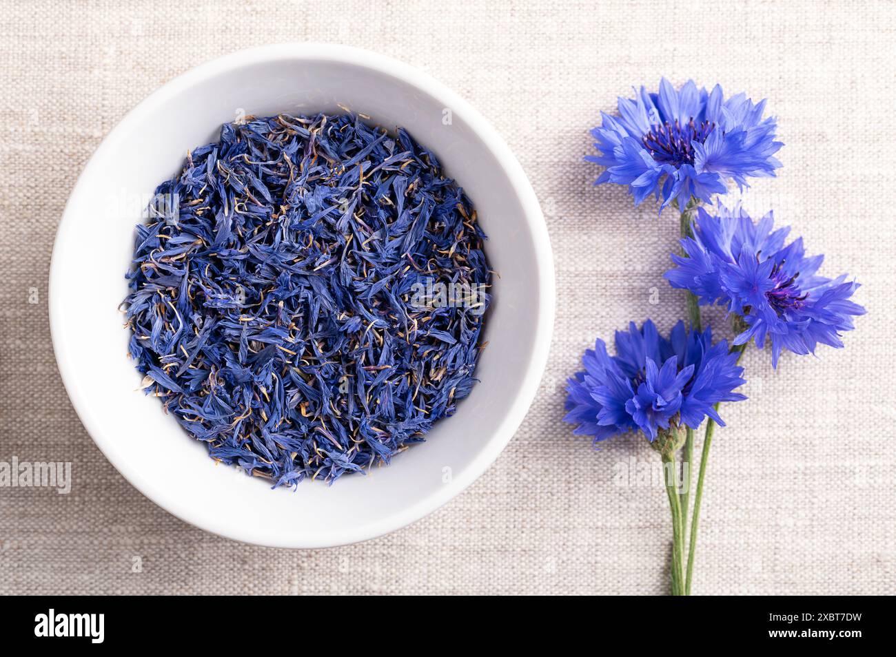 Getrocknete blaue Kornblumenblätter in einer weißen Schüssel auf Leinengewebe. Essbare Blüten von Centaurea cyanus, auch als Junggeselenknopf bekannt. Stockfoto