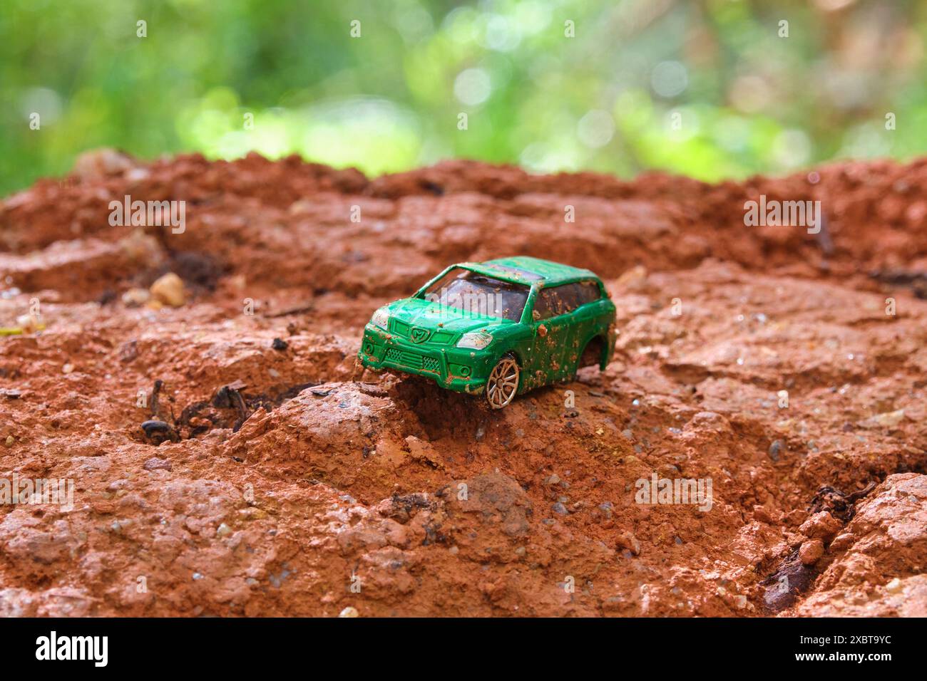 Ein abgenutztes und kaputtes grünes Spielzeugauto mit fehlendem Rad und beschädigter Windschutzscheibe auf einem Hügel aus rötlich-braunem Schlamm mit grünem, traumhaftem Bokeh-Hintergrund. Stockfoto