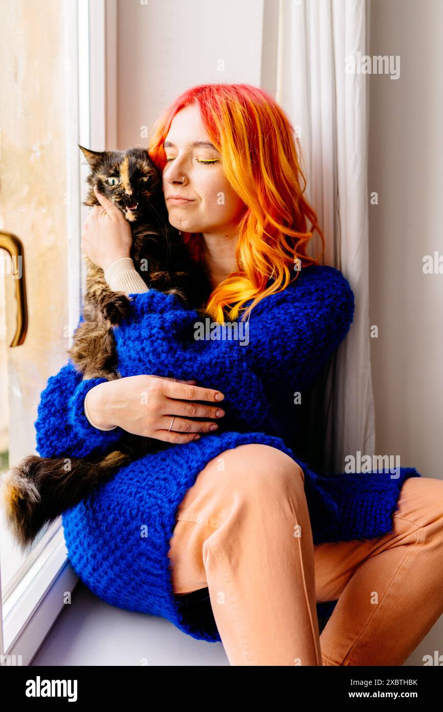 Eine Frau mit orangefarbenen Haaren hält eine Katze im Arm. Die Katze ist schwarz-weiß. Die Frau trägt einen blauen Pullover und eine orange Hose Stockfoto
