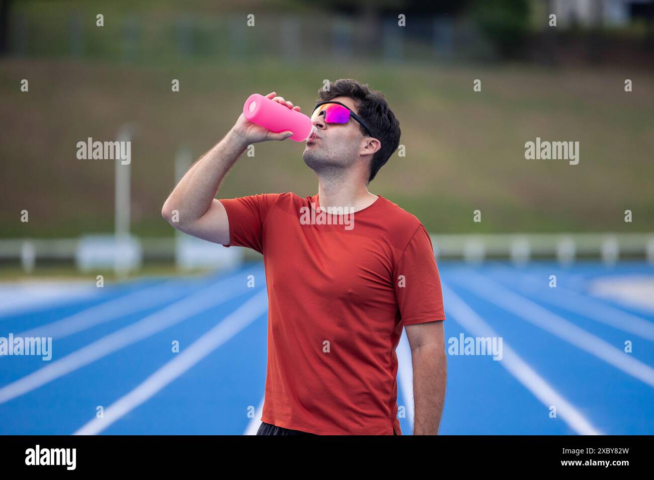 Ein Läufer mit einer Sonnenbrille trinkt Wasser aus einer rosa Flasche, nachdem er sein Rennen auf einer Leichtathletikstrecke beendet hat. Dieses Bild zeigt den Moment von hydra Stockfoto