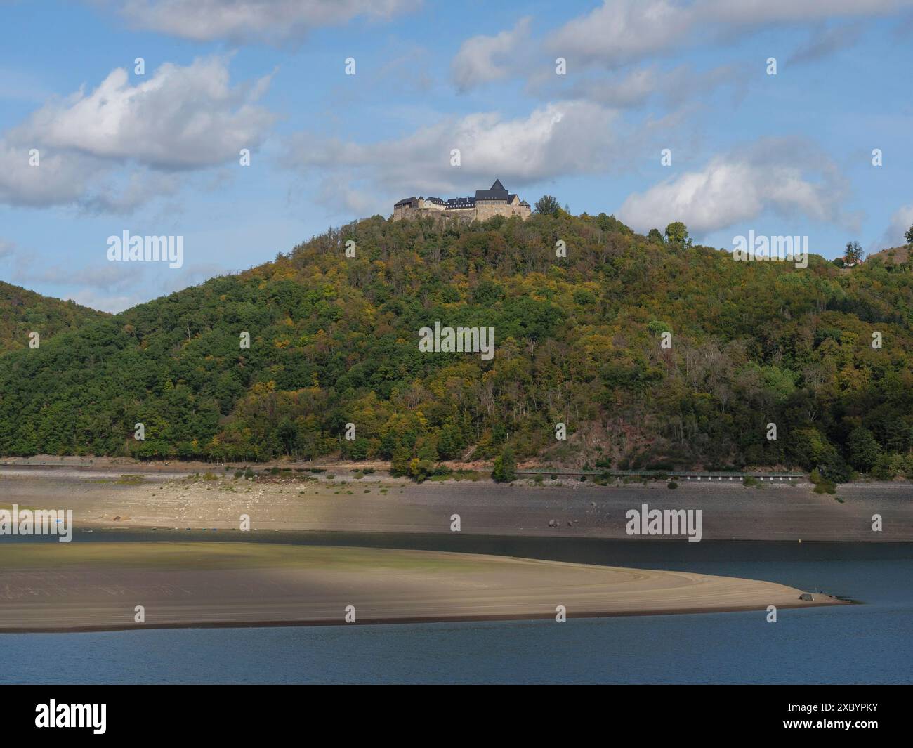 Ein markantes Schloss überblickt die Szene von einem bewaldeten Hügel, der sich hinunter zum Ufer eines Flusses hinabschrägt, unter einem hellen Himmel mit Wolkenformationen, Waldeck Stockfoto