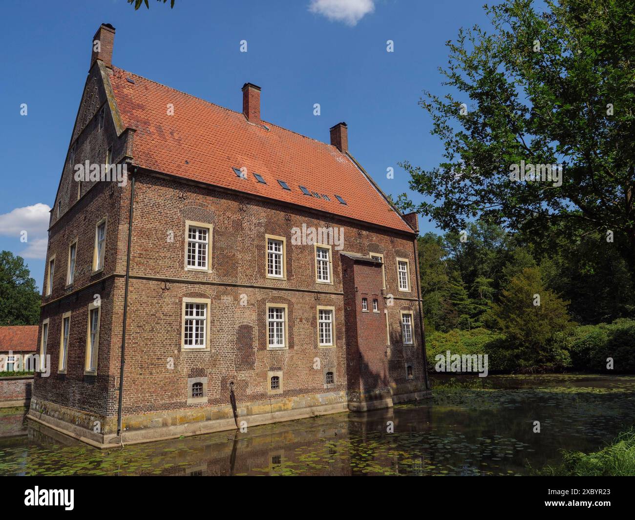 Altes Backsteinhaus mit rotem Dach am Rande eines Teiches, im Sommer von grünen Bäumen umgeben, ochtrup, münsterland, deutschland Stockfoto