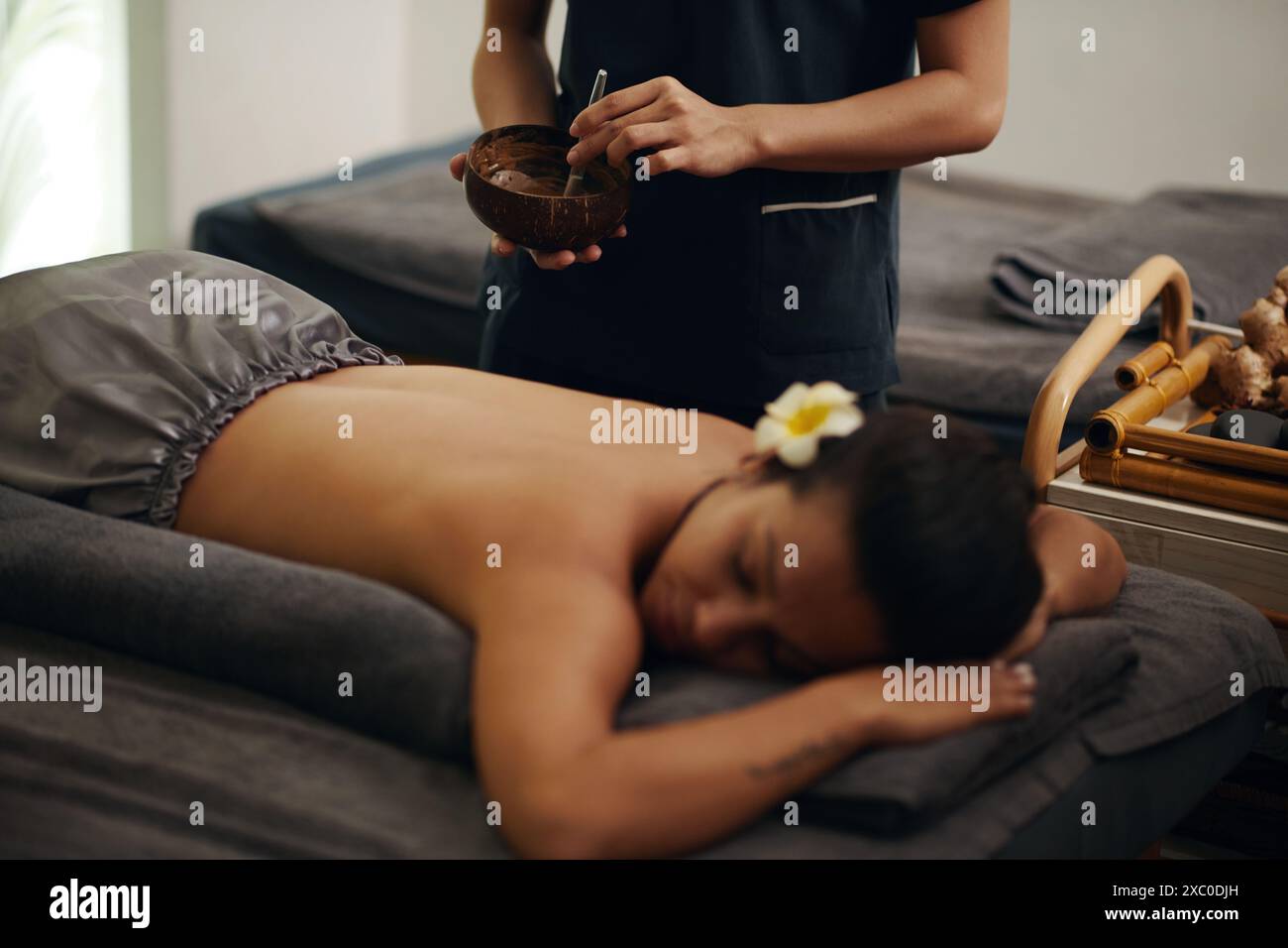 Kosmetikerin mischt Kaffeeschalen in einer Holzschale, bevor sie es auf den Rücken der jungen Frau aufträgt Stockfoto