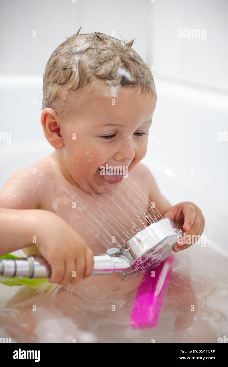 Kleines Kind in einer Badewanne. Glücklicher kleiner Junge genießt in der Badewanne mit Schaumblasen und spielt mit der Dusche. Hygiene und Betreuung von Kleinkindern Stockfoto