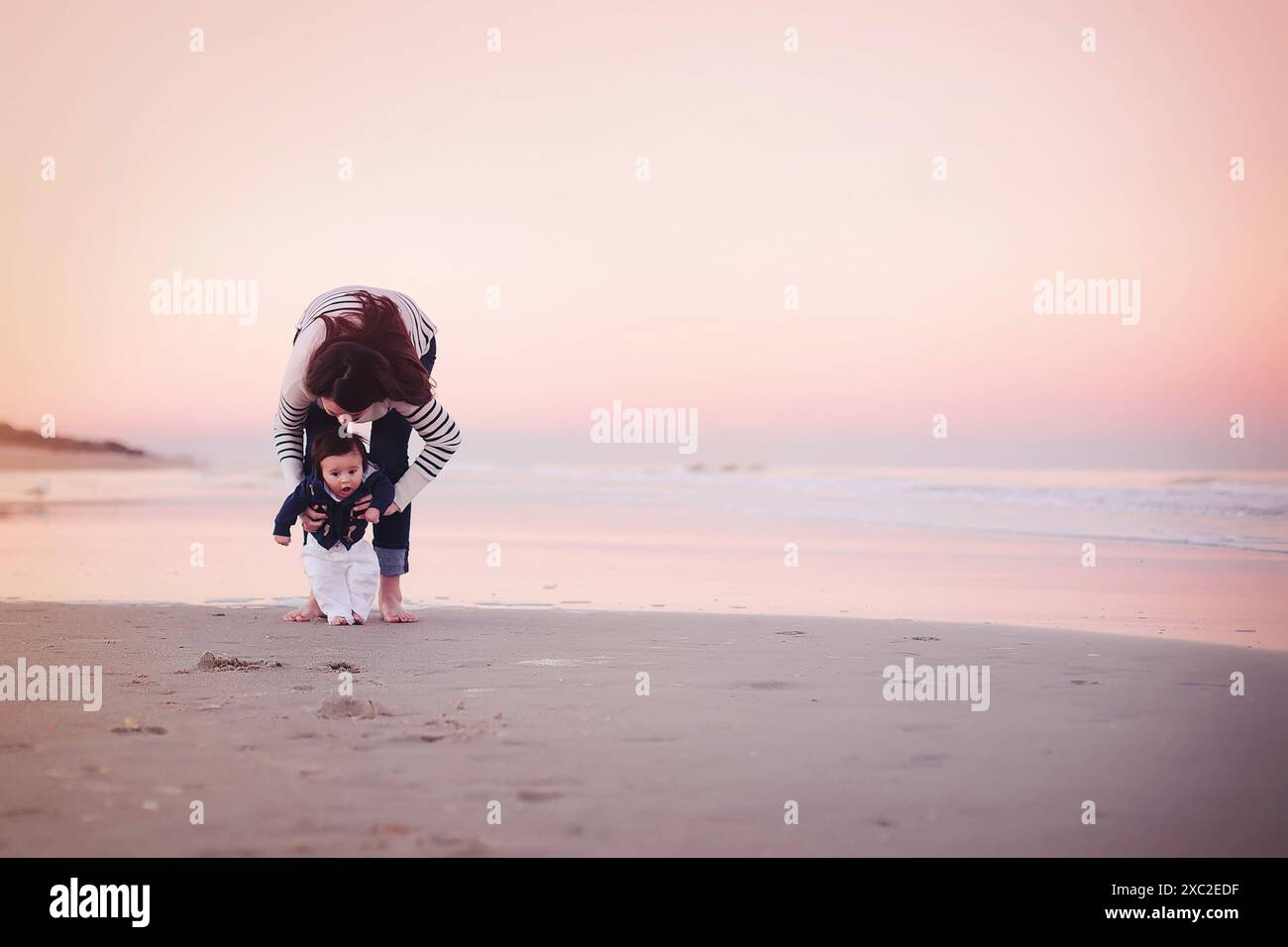 Mutter hält ihr Baby am Strand bei Sonnenuntergang, Baby sucht nach Stockfoto