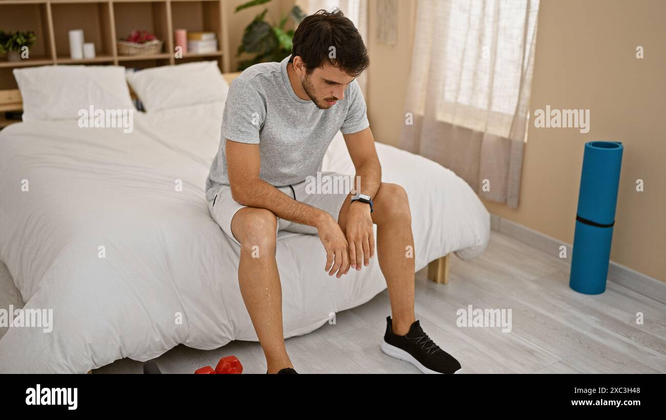 Nachdenklicher junger Mann, der auf einem Bett in einem Schlafzimmer mit Trainingsausrüstung sitzt und über seine Fitness-Routine nachdenkt. Stockfoto