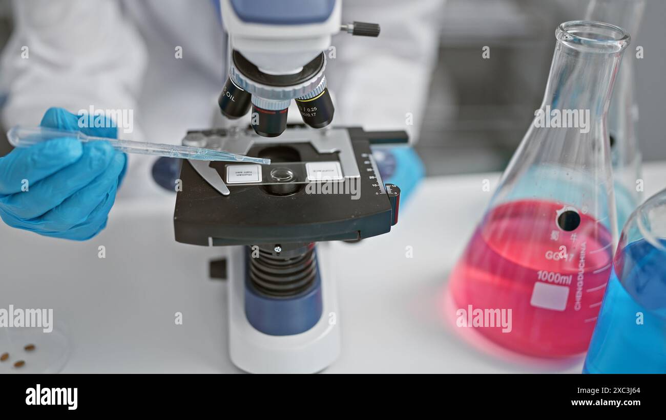 Ein Forscher in Handschuhen verwendet eine Pipette über einem Mikroskop neben farbenfrohen Laborkolben in einem Labor Stockfoto