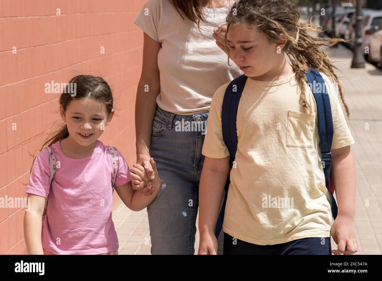Die junge Mutter bringt ihre Kinder zu Beginn des Schuljahres zur Schule. Sie reden und lächeln an einem sonnigen Tag. Zurück zur Schule, Rucksäcke, Familie, am ersten Tag Stockfoto