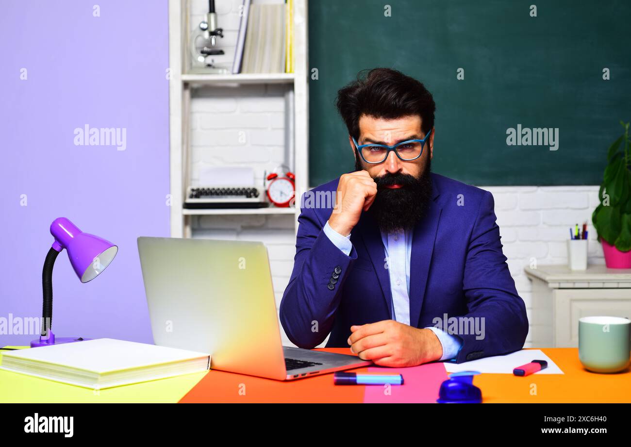 Ernsthafter männlicher Universitätslehrer, der mit Laptop im Klassenzimmer arbeitet. E-Learning und Bildung. Online-Test oder Prüfung im College. Bärtige Studenten Stockfoto