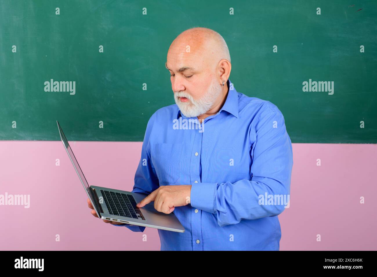 Ernsthafter männlicher Lehrer in lässigem Hemd mit Laptop im Klassenzimmer. Hochschulprüfung. Schullehrer in der Nähe der Tafel, der den Schülern Unterricht gibt Stockfoto