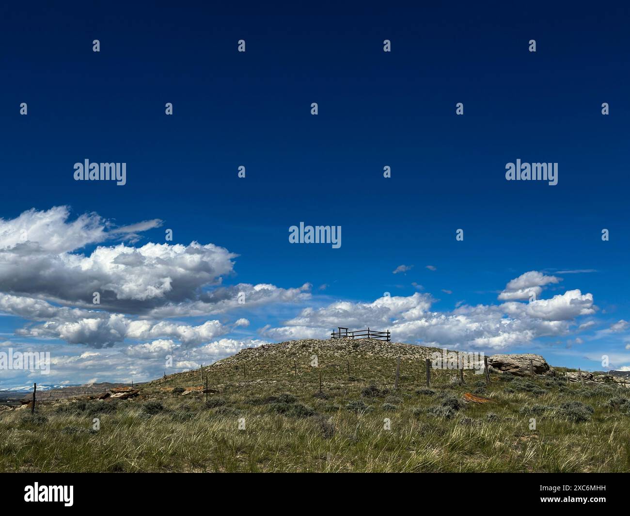 Ein hoher Hügel in Cody, Wyoming, ist von weißen Wolken und blauem Himmel umgeben. Alte Holzzäune umgeben den Hügel mit einem Korral oder einem Stift an der Spitze. Stockfoto