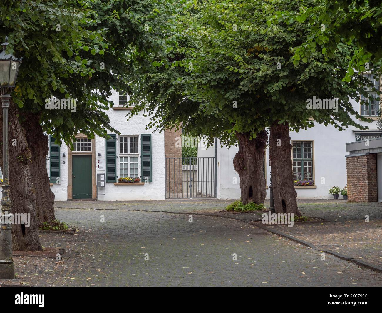 Ruhiger Innenhof mit Kopfsteinpflaster, alten Bäumen und einem weißen Gebäude mit Fensterläden, Xanten, Nordrhein-Westfalen, Deutschland Stockfoto