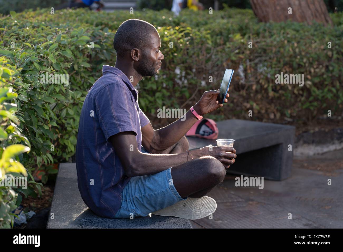 Ein junger Afroamerikaner sitzt draußen in einem Park, benutzt ein Smartphone und hält einen Drink - zwanglose und entspannte Szene - moderne Technik, Freizeit Stockfoto