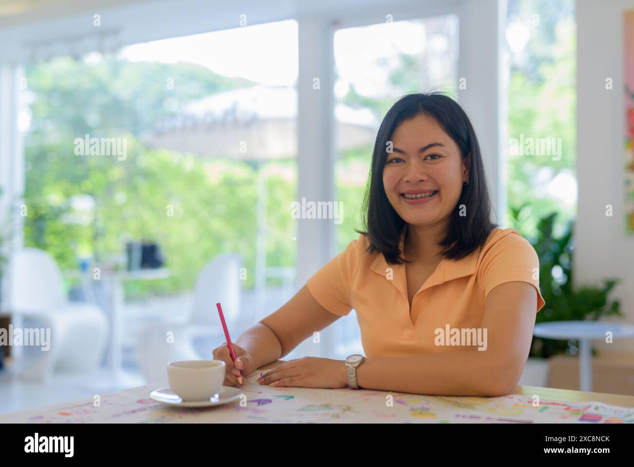 Frau, die im Café sitzt. Sie trägt lässig ein orangefarbenes Oberteil und eine Zeichnung. Stockfoto