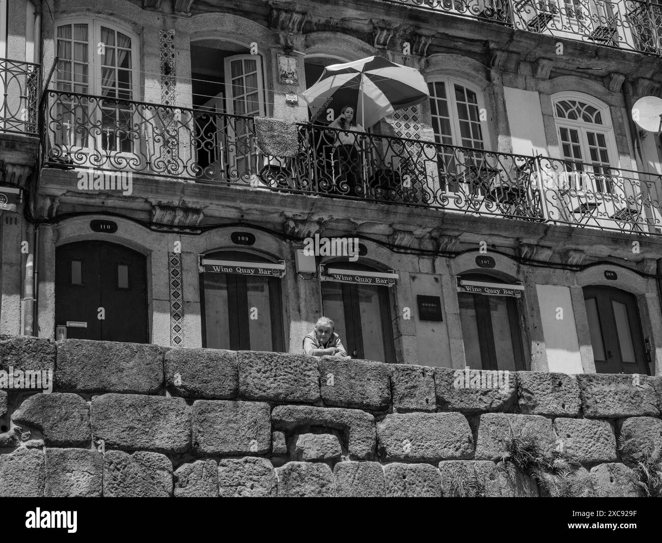 Junge Frau, die auf ihrem Balkon steht, bedeckt unter einem Sonnenschirm, der mit dem Telefon spricht, alte Frau, die auf der Straße Porto beobachtet. Portugal Stockfoto