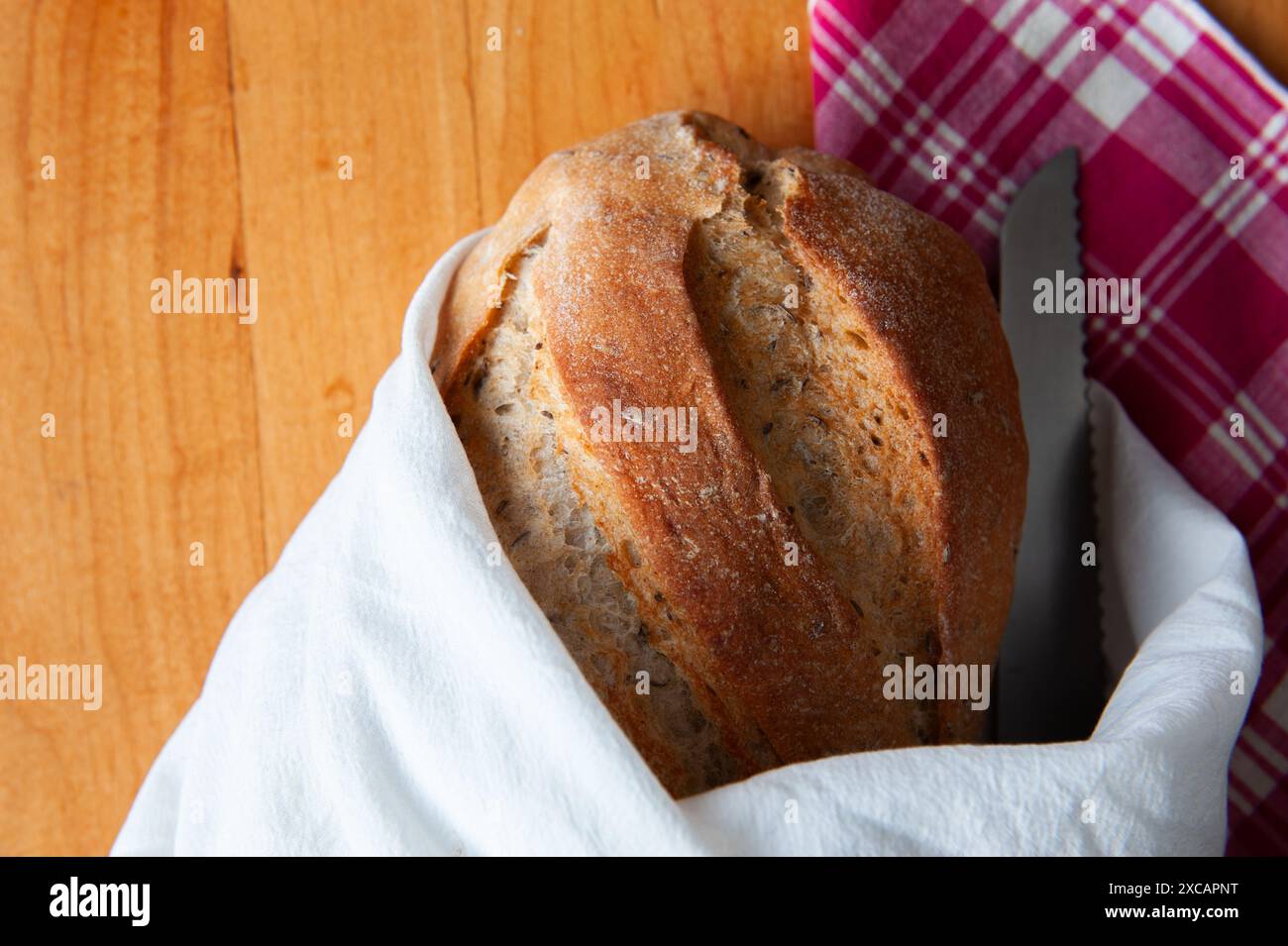 Ein frischer Brotlaib Rosmarin in ein weißes Leinentuch mit einem gezackten Brotmesser auf einem Holztisch. Stockfoto