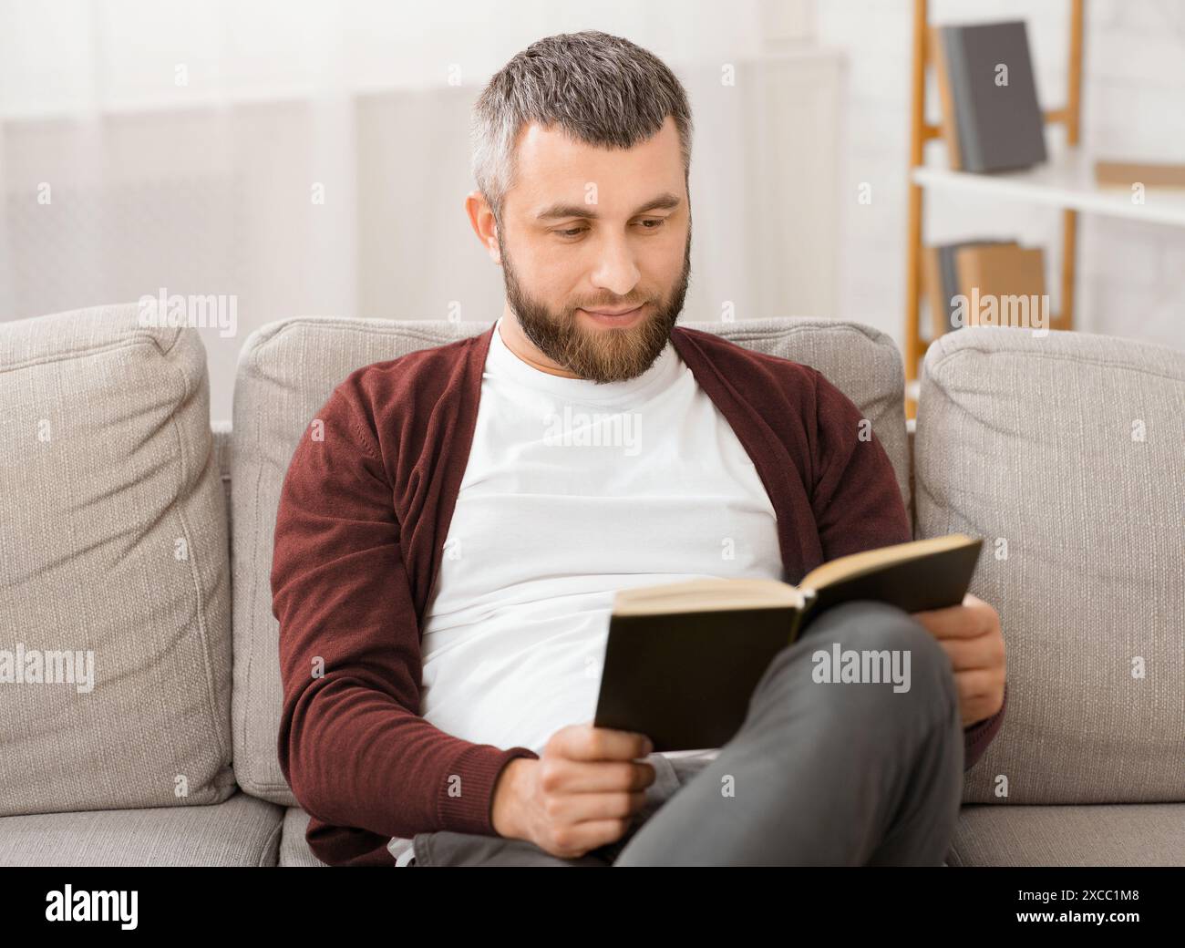 Bärtiger Mann sitzt auf der Couch und liest Buch Stockfoto