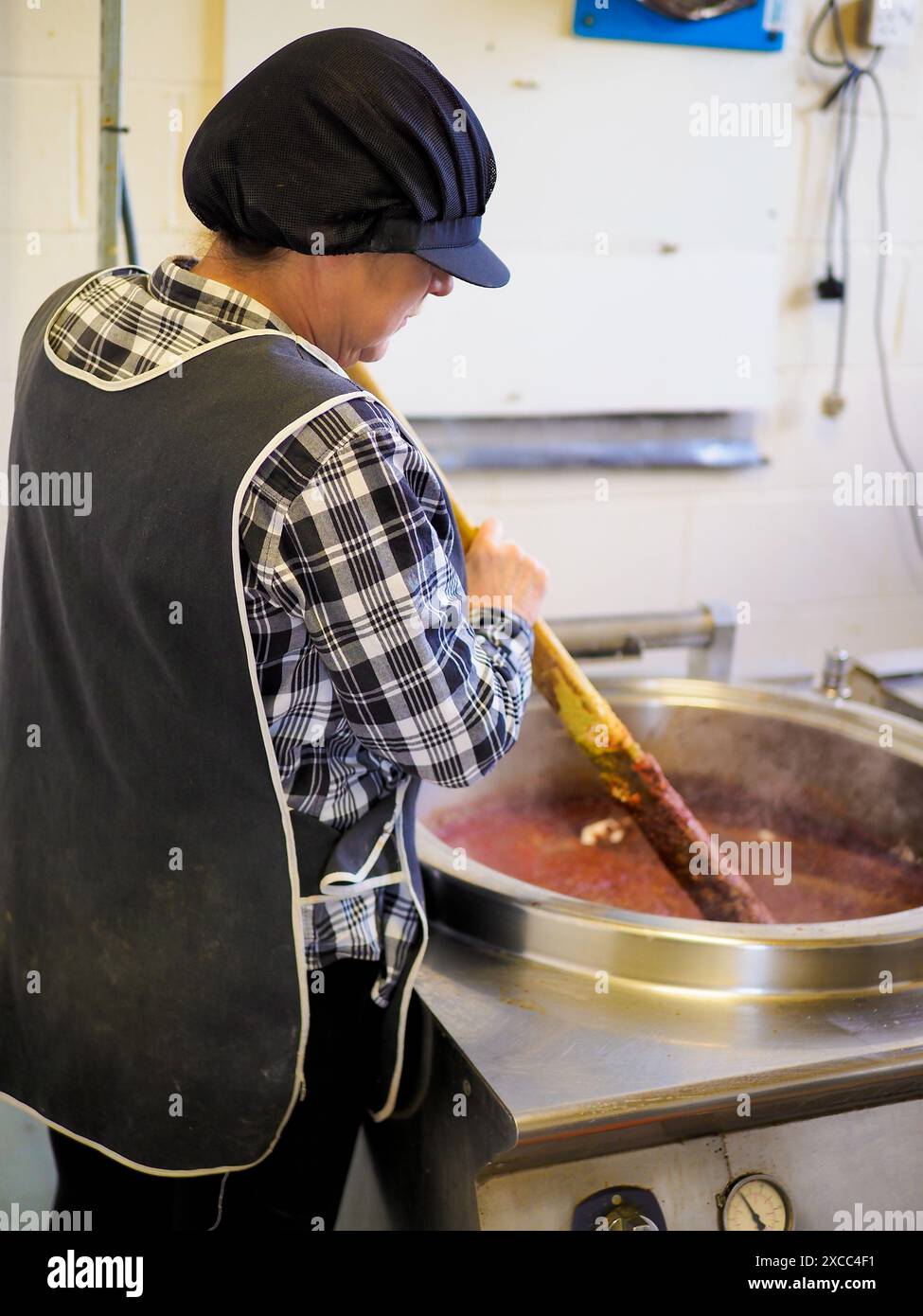 Frau rührt großen Topf in der kommerziellen Küche, trägt kariertes Hemd und schwarze Schürze. Stockfoto