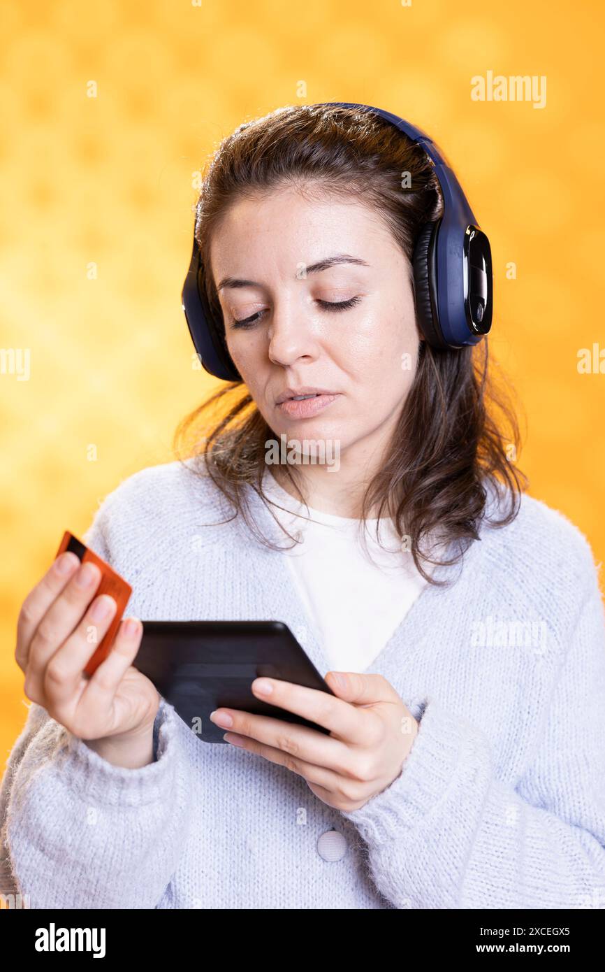 Frau, die elektronische Bücher für ereader mit Kreditkarte kauft, isoliert über Studiohintergrund. Bücherwurm hört Musik in Kopfhörern und kauft E-Books, die mit dem Gerät gelesen werden können Stockfoto