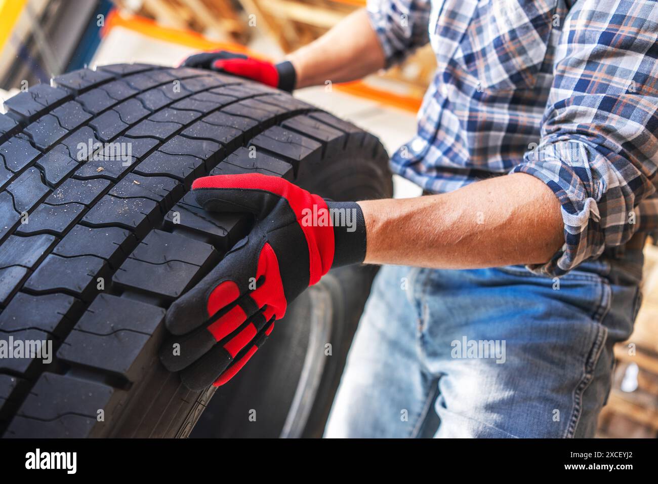 Ein Mechaniker mit kariertem Hemd und Jeans untersucht einen großen Lkw-Reifen mit roten Handschuhen. Er hält den Reifen mit der linken Hand und überprüft Stockfoto