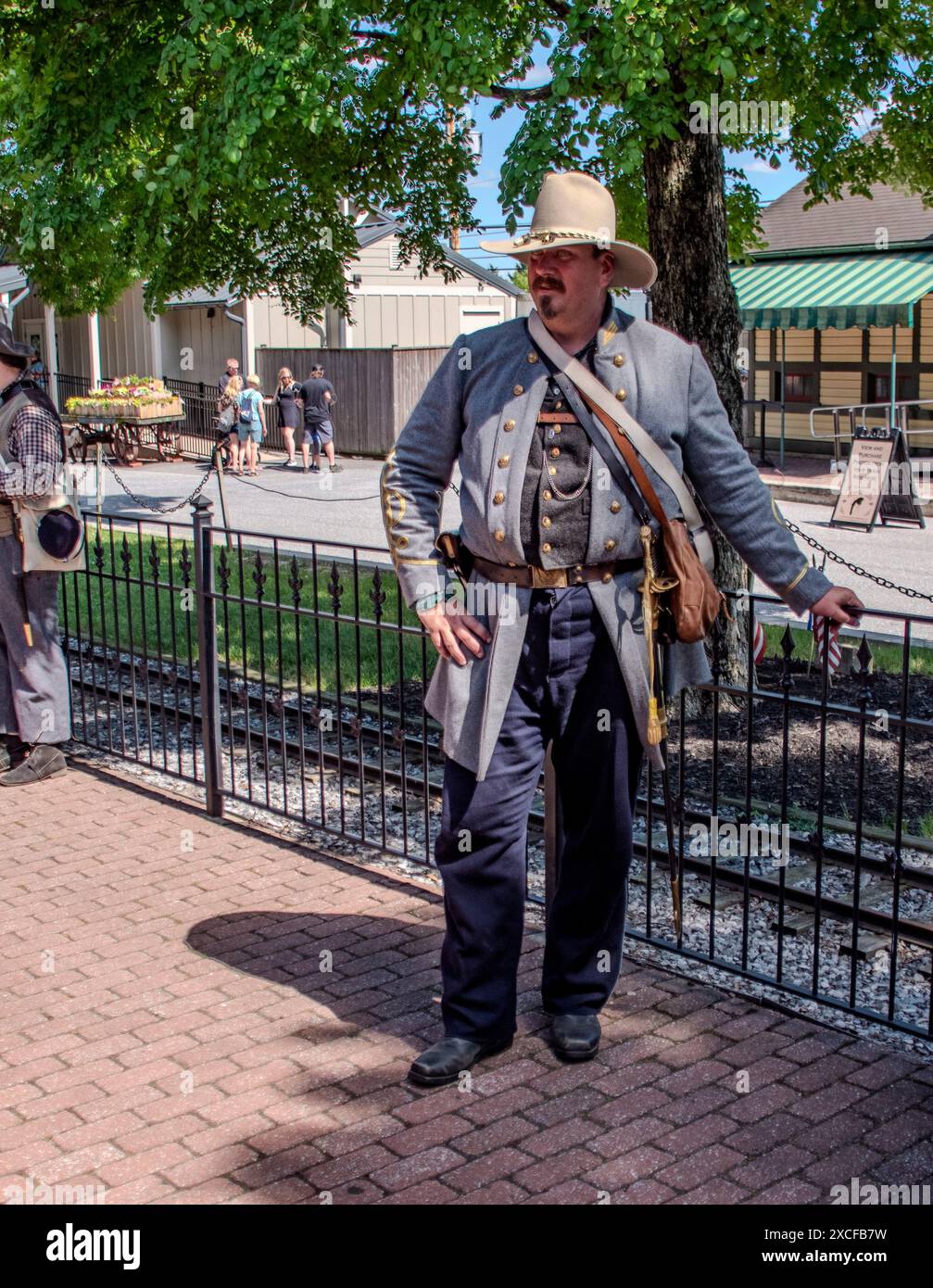 Strasburg, Pennsylvania, USA, 8. Juni 2024 - Ein Reenactor des Bürgerkriegs in Einem grauen Militärmantel mit goldenen Akzenten und Einem Beige Hut steht beiläufig neben Einem Black Iron Fence in der Nähe von Eisenbahngleisen, mit Bäumen und modernen Gebäuden im Hintergrund unter Einem klaren Himmel. Stockfoto