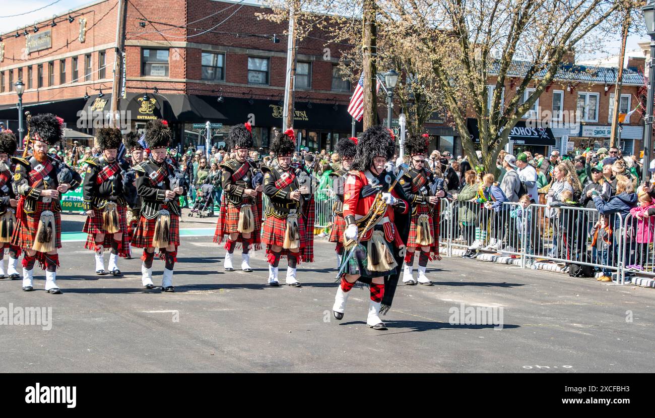 Lindenhurst, New York, USA, 30. März 2024 - Eine Gruppe von Dudelsackern in traditionellen schottischen Kilts und Bärenhauthüten tritt an Einem sonnigen Tag in Einer bunten Straßenparade auf, umgeben von Zuschauern in Einer geschäftigen urbanen Umgebung. Stockfoto