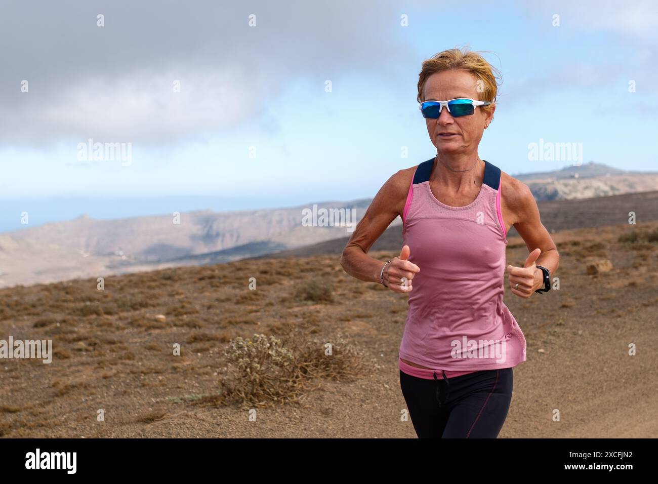 Frau, die draußen in einer trockenen Landschaft mit Bergen im Hintergrund läuft, Sportausrüstung und Sonnenbrille trägt. Stockfoto