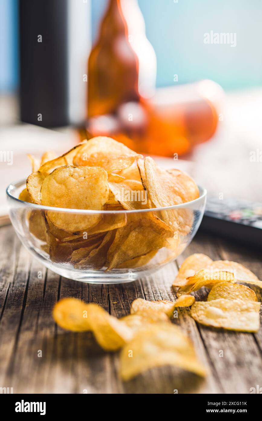 Eine Schüssel mit Kartoffelchips steht auf einem Holztisch, neben einer Fernbedienung und zwei Bierflaschen. Der Fokus liegt auf der Schüssel mit Chips, die A Stockfoto