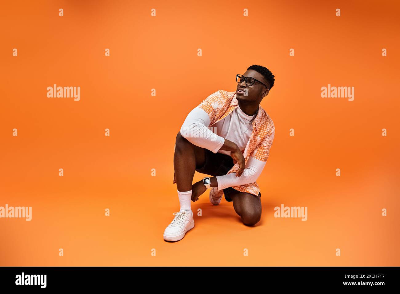 Modischer afroamerikanischer Mann, der sich auf leuchtendem orangefarbenem Hintergrund hockt. Stockfoto