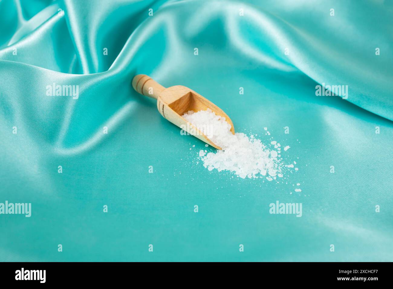 Hölzerne Schaufel, die weißes Steinsalz verschüttet, auf türkisfarbenem Satin-Hintergrund, Nahaufnahme Stockfoto