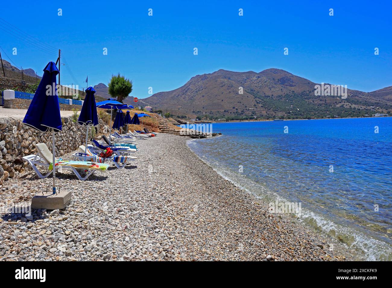 Verlassene Liegestühle und Sonnenschirme an einem steinigen Strand, Tilos Island, Dodekanese, Griechenland. Vom Mai 2024 Stockfoto