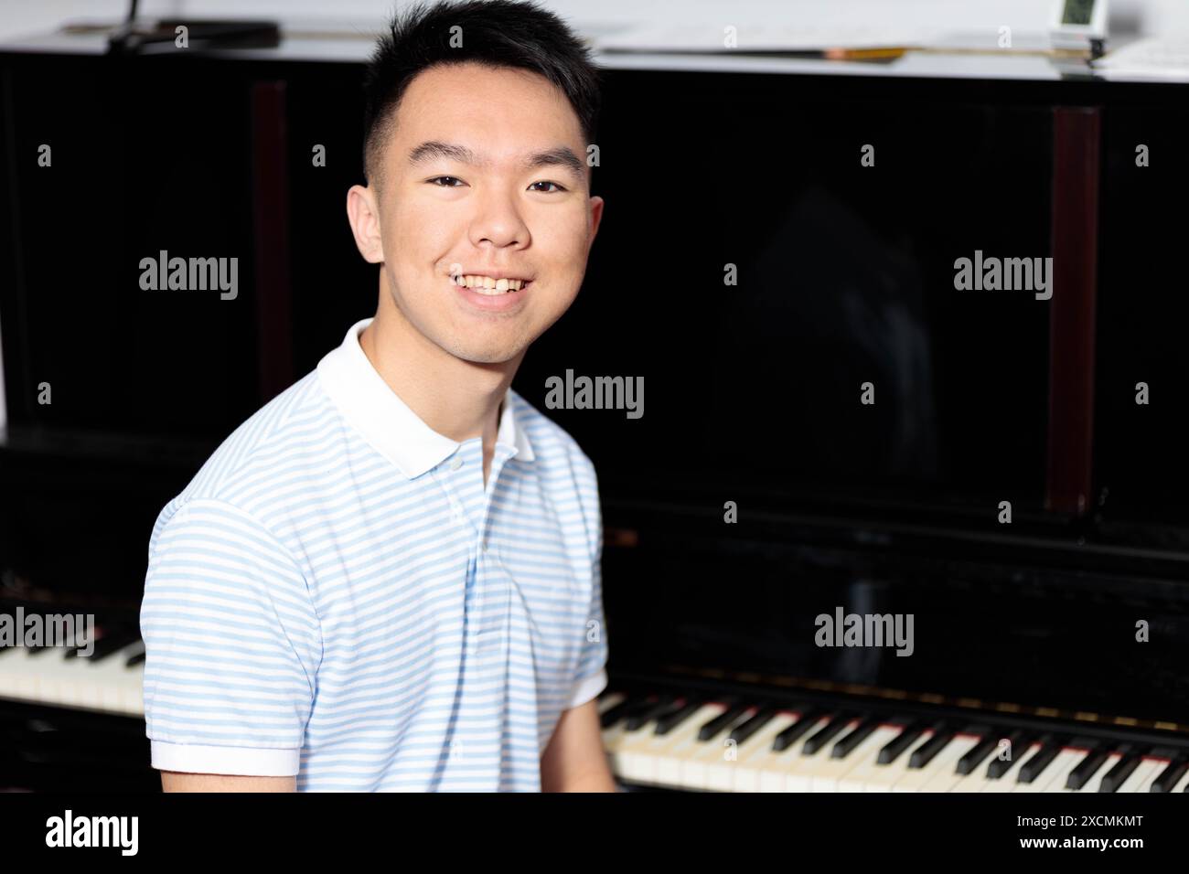 Ein asiatischer Teenager sitzt glücklich an einem Klavier in seinem Haus und blickt direkt in die Kamera mit einem Lächeln im Gesicht, um ein Porträt zu erhalten. Stockfoto