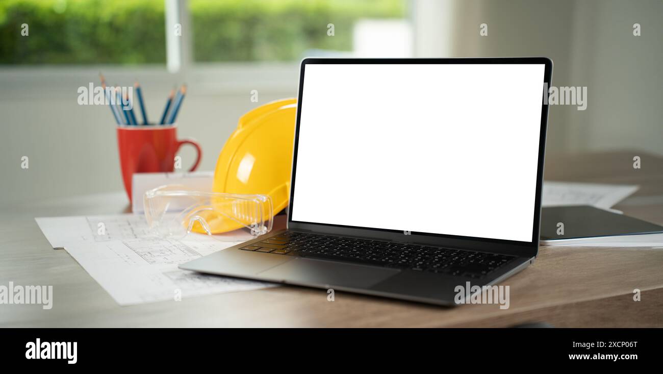 Tischarchitektur-Blaupausen und Hardhat-Laptop auf einem Schreibtisch in einer Büroumgebung, mit einem leeren Bildschirm, der Technologie und Arbeit darstellt Stockfoto