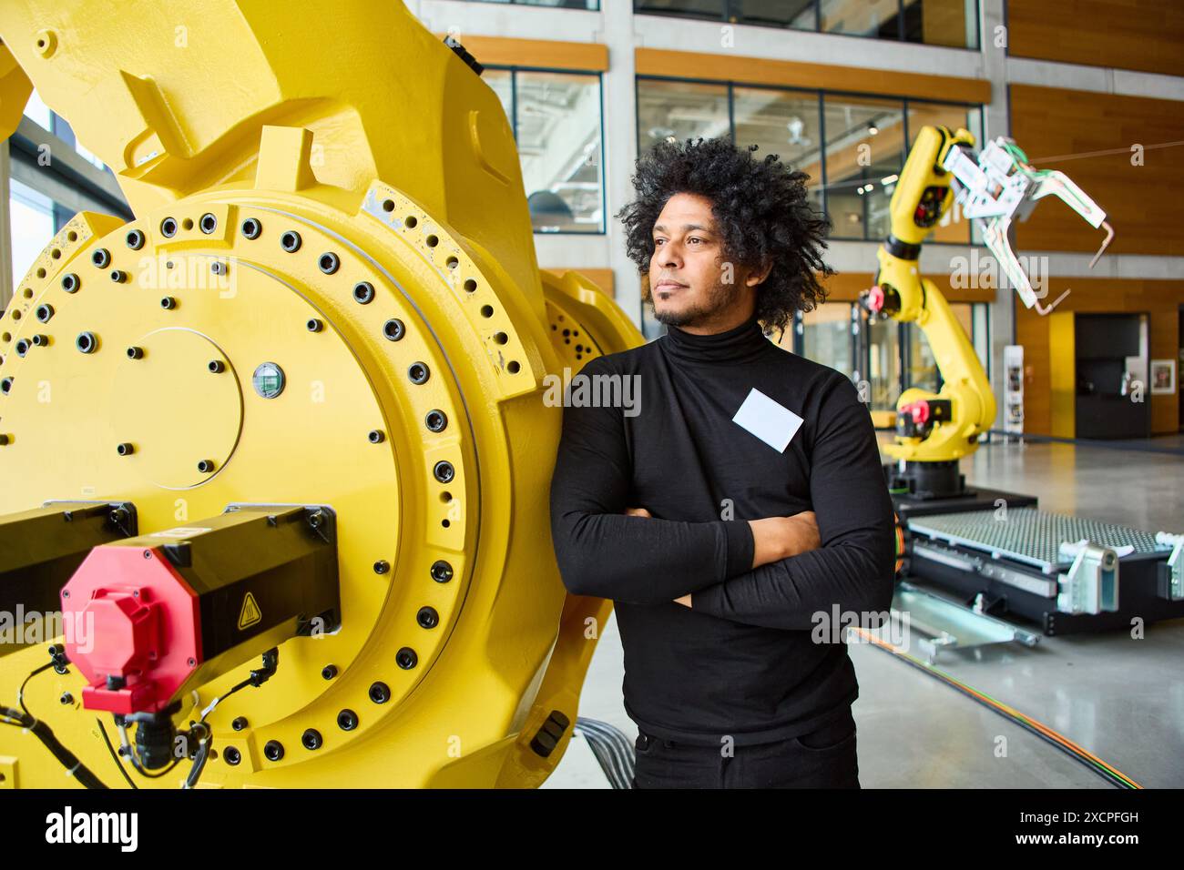 Der Ingenieur steht selbstbewusst mit gekreuzten Armen neben einem großen Roboterarm und präsentiert Industrierobotik und Automatisierung in einem modernen Arbeitsbereich. Stockfoto