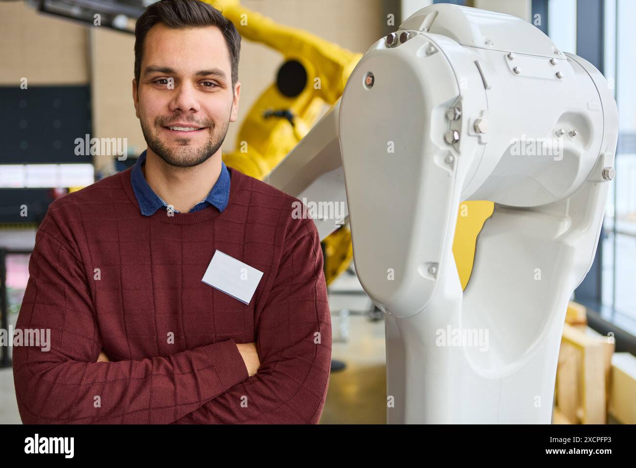 Ein Ingenieur posiert selbstbewusst neben dem weißen Roboterarm in einem modernen industriellen Umfeld und hebt die Robotik und Prozessautomatisierung hervor. Stockfoto