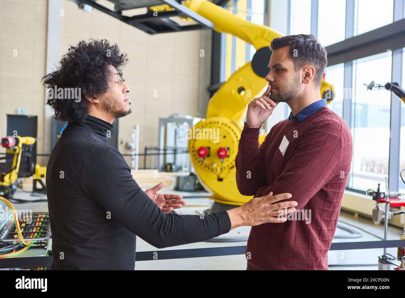 Zwei Ingenieure diskutieren intensiv über Robotik-Technologie, die in einer industriellen Umgebung mit einem gelben Roboterarm im Hintergrund stehen. Stockfoto