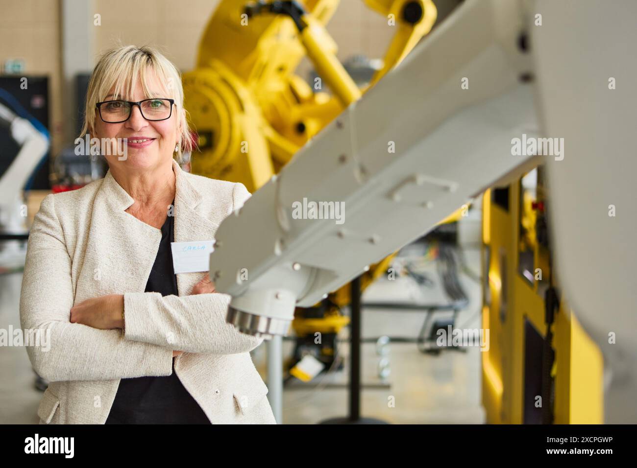 Selbstbewusste Ingenieurin, die mit gekreuzten Armen neben Robotergeräten in einer industriellen Umgebung steht. Konzept von Automatisierung, Technologie und Indu Stockfoto