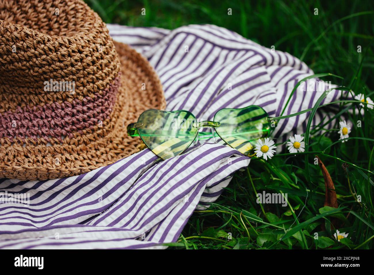 Grüne Sonnenbrille in Herzform und Strohhut auf einem gestreiften Tuch auf einer grünen blühenden Wiese mit weißen Gänseblümchen. Stillleben im Sommer. Sommerurlaub Stockfoto