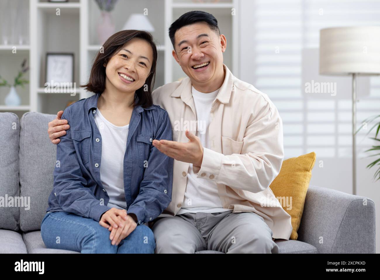 Junge asiatische Paare Mann und Frau, die zu Hause auf der Couch vor der Kamera sitzen und online chatten, stellt der Kerl seine Freundin vor und zeigt mit seiner Hand auf sie. Stockfoto