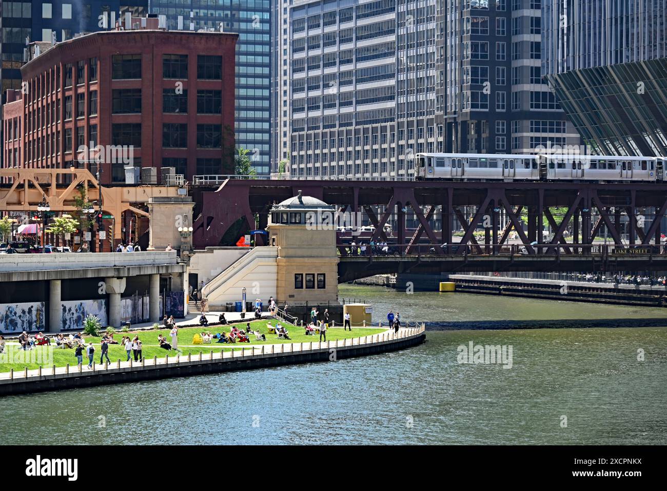 Die Menschen entspannen sich auf dem grasbewachsenen Abschnitt des Riverwalk in der Nähe der Lake Street Bridge, wo sich die drei Zweige des Chicago River in der Innenstadt kreuzen. Stockfoto