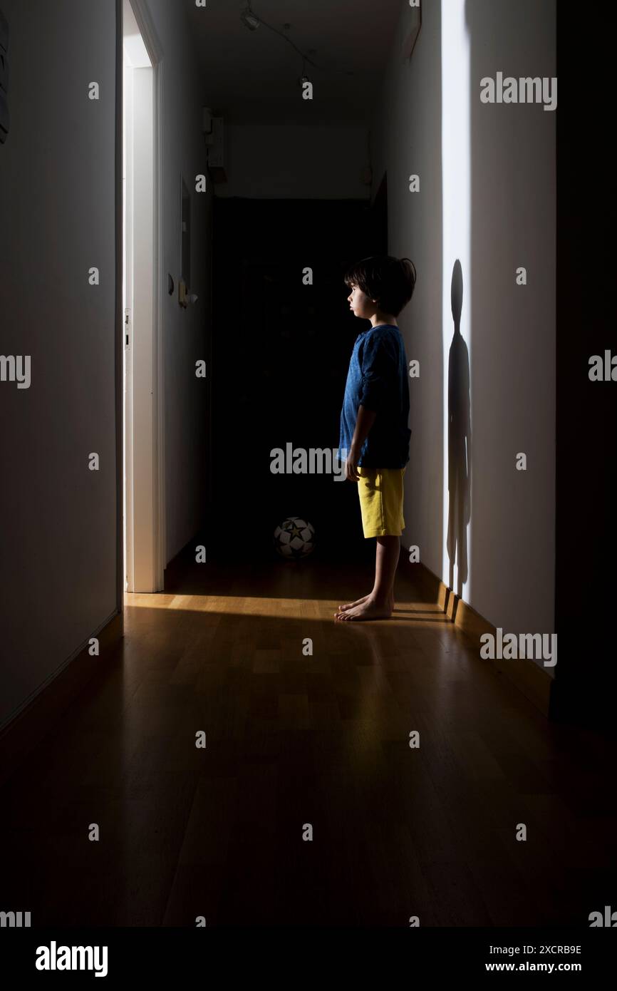 Ein Kind steht in einem dunklen Flur, seine Silhouette wird von einem Lichtstrahl aus einer offenen Tür lang an die Wand gegossen. Stockfoto