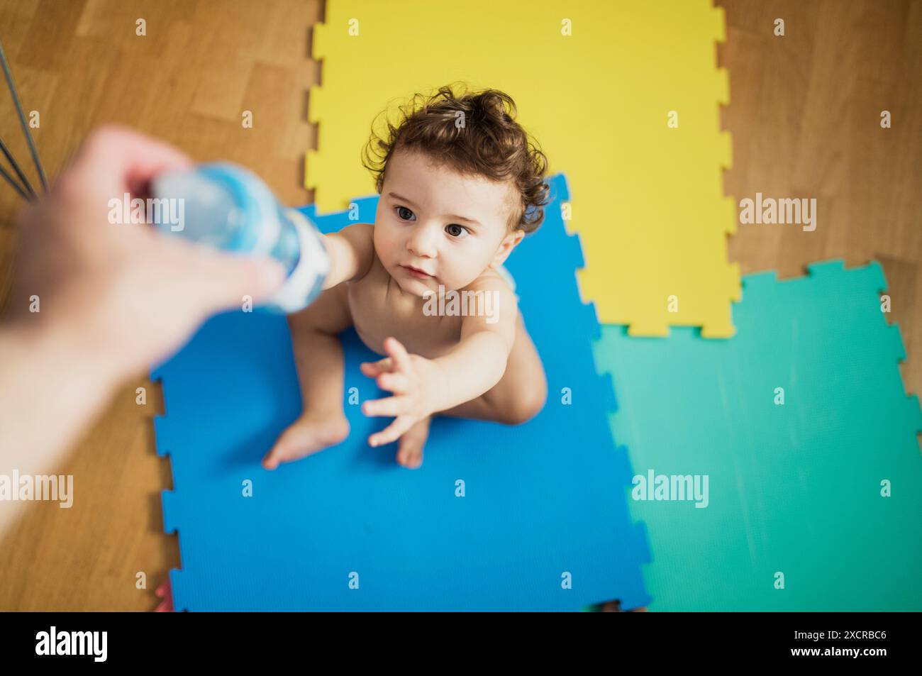 Ein kleiner Junge sitzt auf einem blau-gelben Playmat und greift mit neugierigem Ausdruck auf eine Flasche, die von einer ausgestreckten Hand gehalten wird. Stockfoto