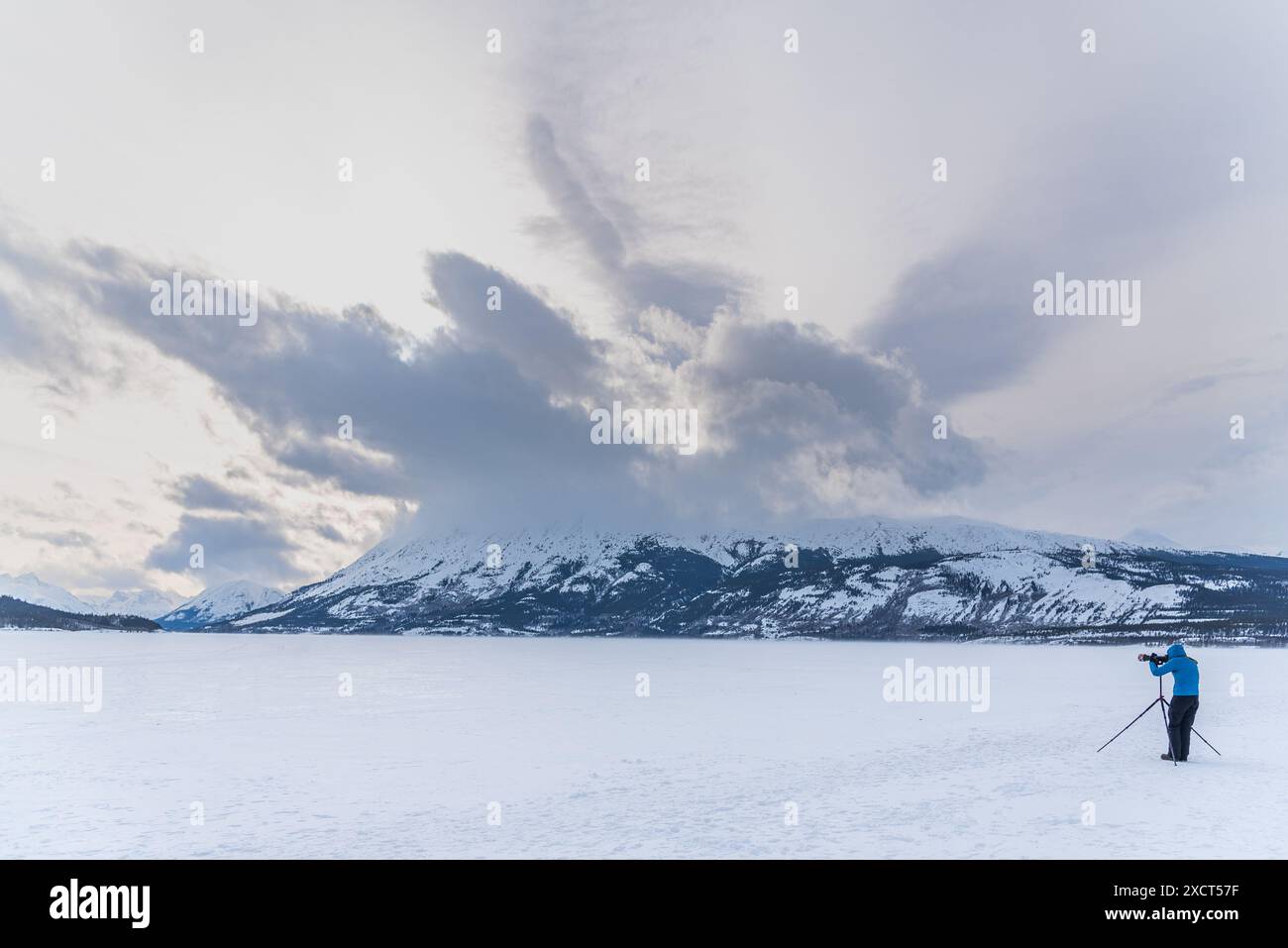 Mann in einer blauen Jacke, der auf einem gefrorenen See steht und Fotos von der atemberaubenden, verschneiten Winterlandschaft mit Wolken über ihnen macht. Stockfoto