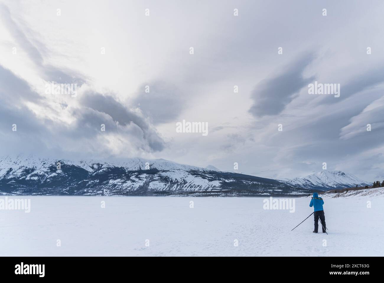 Mann in einer blauen Jacke, der auf einem gefrorenen See steht und Fotos von der atemberaubenden, verschneiten Winterlandschaft mit Wolken über ihnen macht. Stockfoto