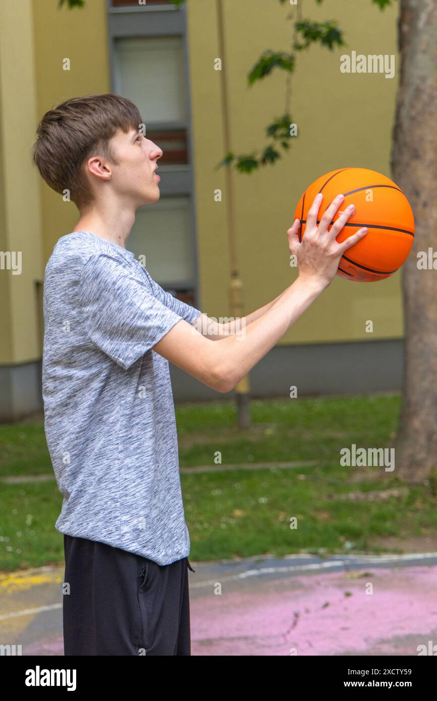 Ein junger Mann, der ein graues T-Shirt und eine schwarze Hose trägt, übt draußen Basketball. Er hält den Ball mit beiden Händen vor der Brust, PR Stockfoto