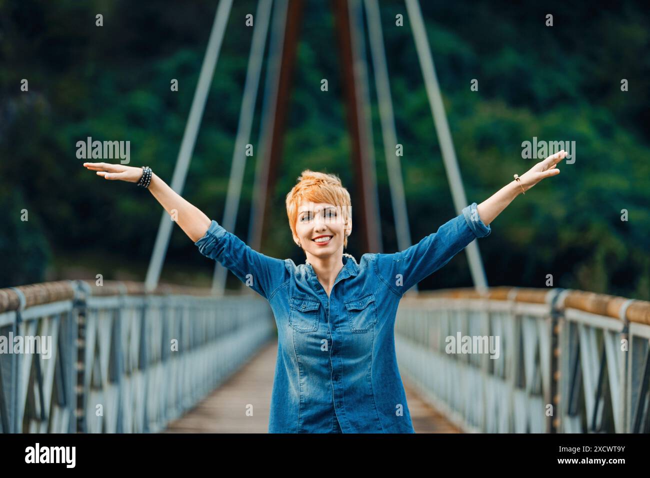 Eine junge Frau mit kurzen roten Haaren steht mit weit geöffneten Armen auf einer Brücke und genießt das Gefühl von Freiheit und Glück. Sie trägt einen Denim Stockfoto