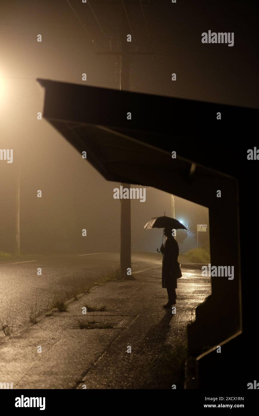 Eine Silhouette einer Frau unter einer Straßenlaterne, die einen Schirm hält, der neben einer alten Bushaltestelle bei Nebel und Regen in Katoomba bei Nacht steht Stockfoto