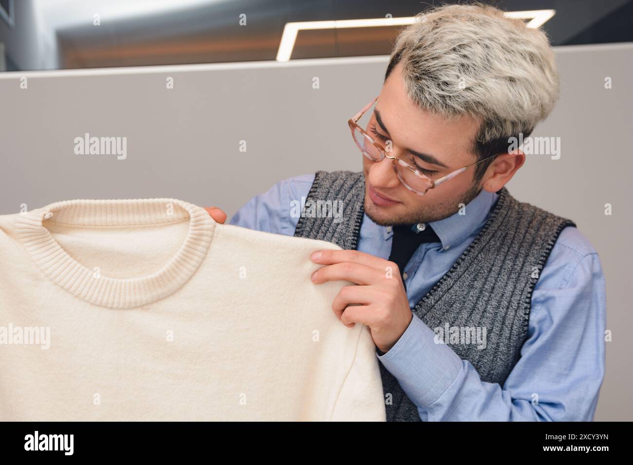 Ein junger Mann in Brille arbeitet, hält und bewertet einen weißen Pullover. Er schaut den Stoff mit einem nachdenklichen Ausdruck an Stockfoto